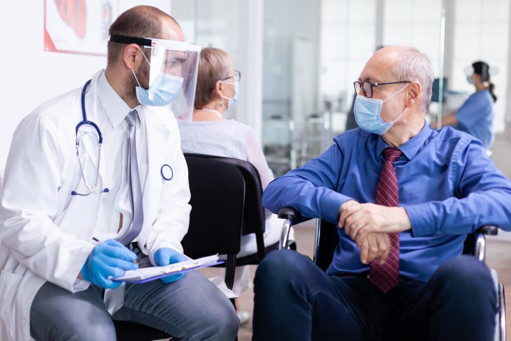 invalid-old-man-with-face-mask-against-infection-with-coronavirus-wheelchair-discussing-with-doctor-hospital-waiting-area